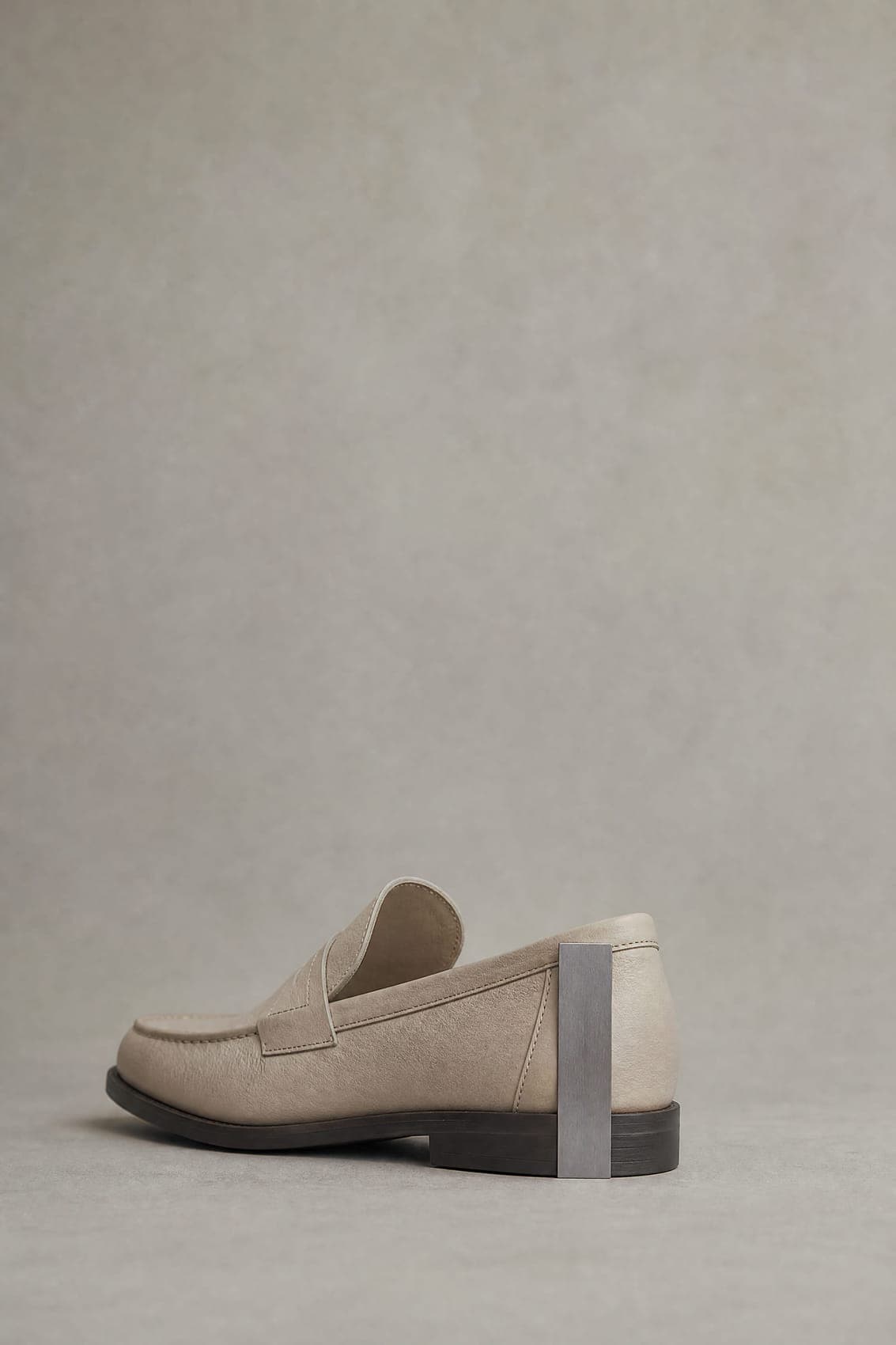 A plain loafer standing on a flat surface, photographed from behind, with an upright gauge blade held against the outside of the heel reaching from the surface to the lowest point of the shoe opening.